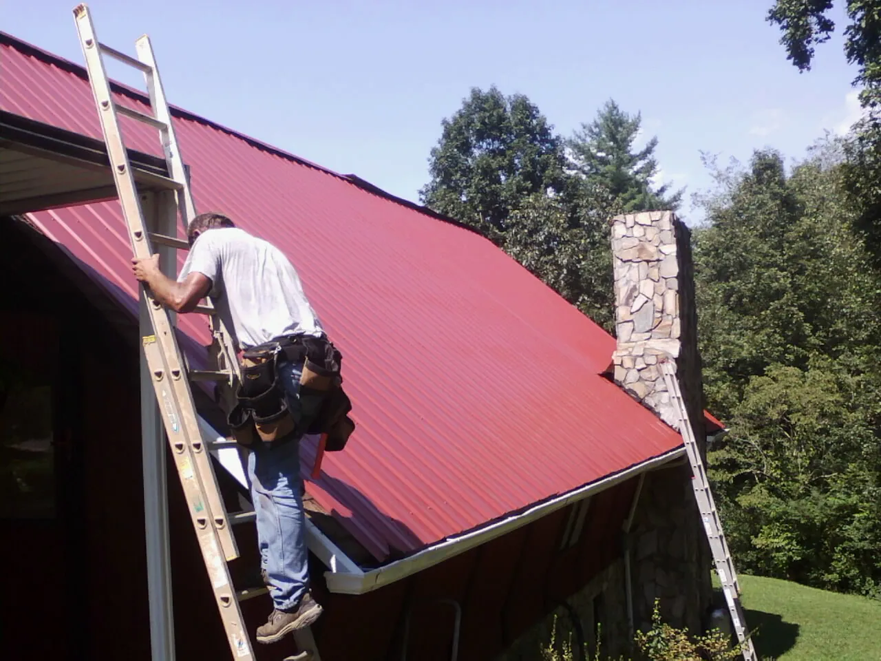 Roofer on a ladder beside a red metal roof and stone chimney — Soffit & Fascia Repair by Roofing Brothers in Cope
